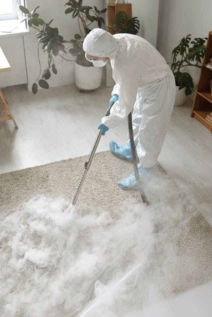 A professional cleaner from Cleaners SW16, dressed in full protective gear including a white suit, gloves, mask, and shoe covers, performs deep cleaning on a beige carpet in a well-lit living room. The cleaner uses a carpet cleaning machine, with visible steam or cleaning solution being applied to the surface. The room features a wooden floor transition, a potted plant, and a modern, minimalist decor with light-colored walls and furniture, demonstrating thorough surface cleaning and sanitisation. The scene illustrates effective deep cleaning practices aimed at maintaining hygiene and freshening residential carpets, aligned with services promoted on the page for avoiding hidden charges in Streatham carpet cleaning quotes, SW16.