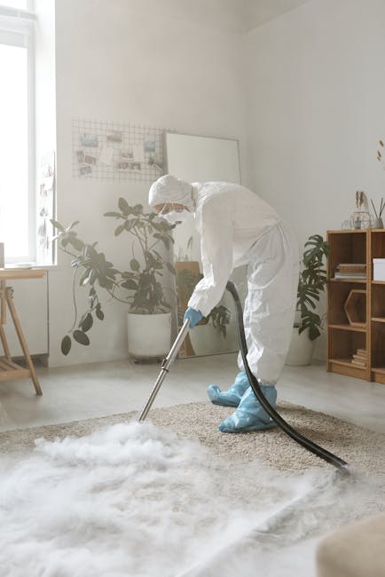 A professional cleaner dressed in a white protective suit, gloves, face mask, and shoe covers is performing deep carpet cleaning in a bright, modern living room. They are using a vacuum or extraction machine with a large black hose, applying it to a light-colored, textured carpet. The room features white walls, a wooden sideboard with decorative items, potted plants near the window, and natural daylight streaming in, creating a clean, fresh atmosphere. The scene illustrates surface cleaning and sanitisation as part of residential or commercial cleaning services provided by Cleaners SW16, emphasizing thorough hygiene and maintenance of indoor surfaces aligned with the page title, 'Avoid hidden charges Streatham carpet cleaning quotes, SW16'.