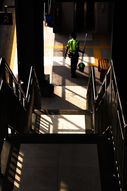 A commercial cleaning staff member from Cleaners SW16 is captured walking down a sunlit staircase inside a railway station, holding a mop and a green bucket. The staircase features metal railings on both sides, casting shadows on the steps. The surrounding area includes tiled floors and station signage in the background, illuminated by natural light streaming through large windows, highlighting the cleanliness and maintained condition of the space. The scene emphasizes professional deep cleaning and surface sanitisation within a busy public transport environment as part of the services offered by Cleaners SW16 in Streatham near Streatham Hill station, SW16.