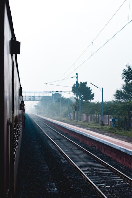 The image depicts a railway station platform with a set of railway tracks running parallel to it. The platform surface appears to be paved with concrete, clean and free of debris. Overhead electrical wiring runs above the tracks, supported by blue metal poles at regular intervals. In the background, there are trees and a partly cloudy sky. The scene is well-lit by natural daylight, and the overall appearance suggests a well-maintained outdoor environment. This setting aligns with the type of location where Cleaners SW16 offers deep cleaning and surface cleaning services near Streatham Hill station, SW16, emphasizing cleanliness and maintenance of public transport infrastructure.