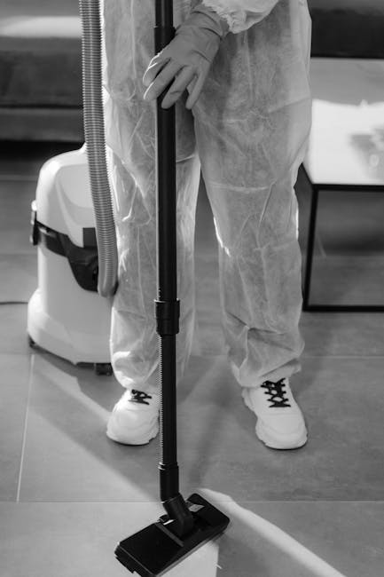 A professional cleaner from Cleaners SW16 is dressed in protective clothing, including gloves and a disposable suit, while using a vacuum cleaner equipped with a floor nozzle for surface cleaning. The cleaner is standing on a polished wooden or laminate floor, cleaning a flat surface area. The scene features a nearby cleaning machine with a flexible hose, indicating a thorough deep cleaning process. The room has good lighting, highlighting the cleanliness and shine of the floor, and the environment appears organized and hygienic, consistent with domestic or commercial surface maintenance. The image visually emphasizes the importance of detailed surface cleaning as part of comprehensive end-of-tenancy cleaning, aligning with the service offerings of Cleaners SW16 and the page’s focus on end of tenancy cleaning price guide in SW16.