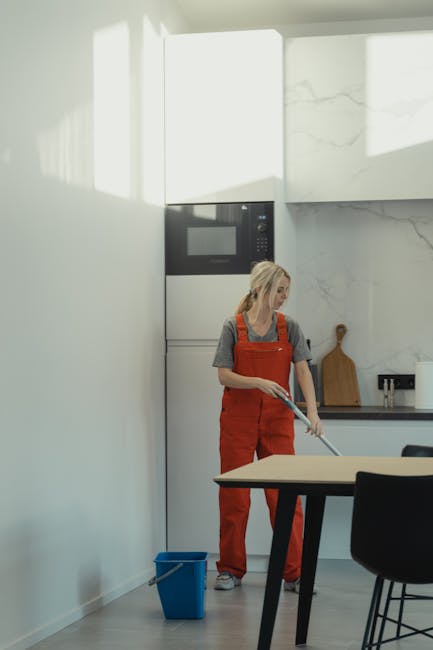 A woman dressed in a grey t-shirt and orange overalls is engaged in surface cleaning with a mop in a modern kitchen. The room features a white wall with a large marbled backsplash and cabinetry, and is illuminated by natural light streaming in from a window or skylight. Visible objects include a small blue bucket on the floor, a wooden cutting board on the countertop, and a black chair partially visible in the foreground. The sleek surfaces appear clean and dry, indicating a thorough deep cleaning process by [COMPANY_NAME] as part of domestic cleaning services for end of tenancy hygiene and sanitisation. The overall scene emphasizes meticulous surface cleaning and maintenance in a contemporary residential setting.